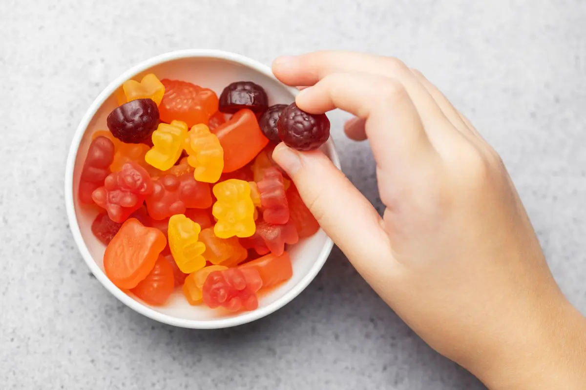 GettyImages-1831831117 A hand reaches for a red gummy candy from a white bowl filled with assorted colorful gummy candies on a gray surface.