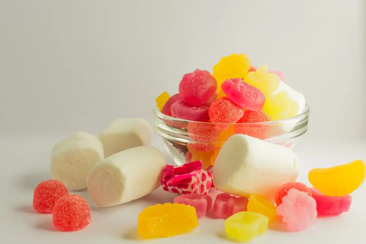 A small glass bowl filled with colorful gummy candies and marshmallows, with additional marshmallows and gummy candies scattered in the foreground.