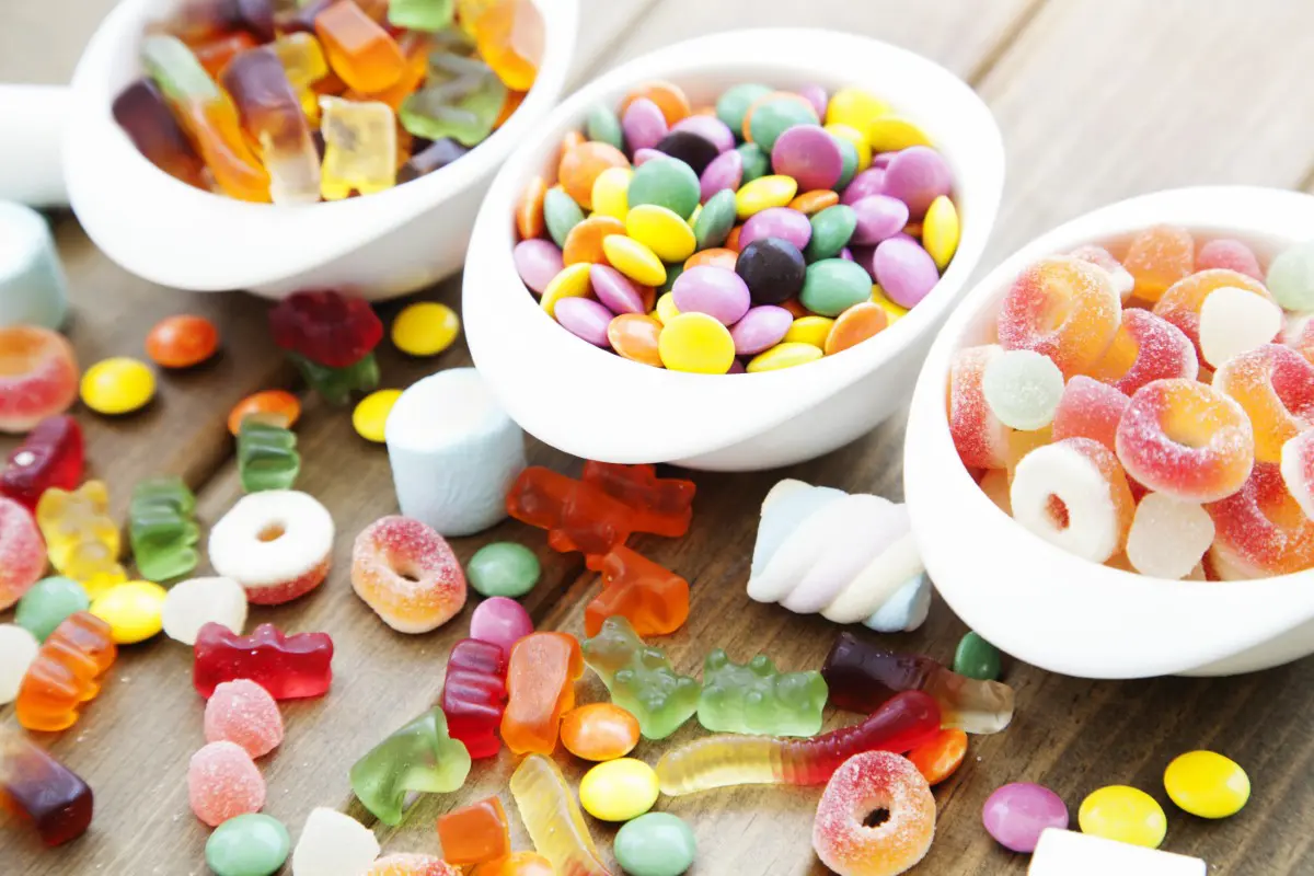 Various colorful candies, including gummy bears, jelly rings, and chocolates, are displayed in white bowls on a wooden surface.